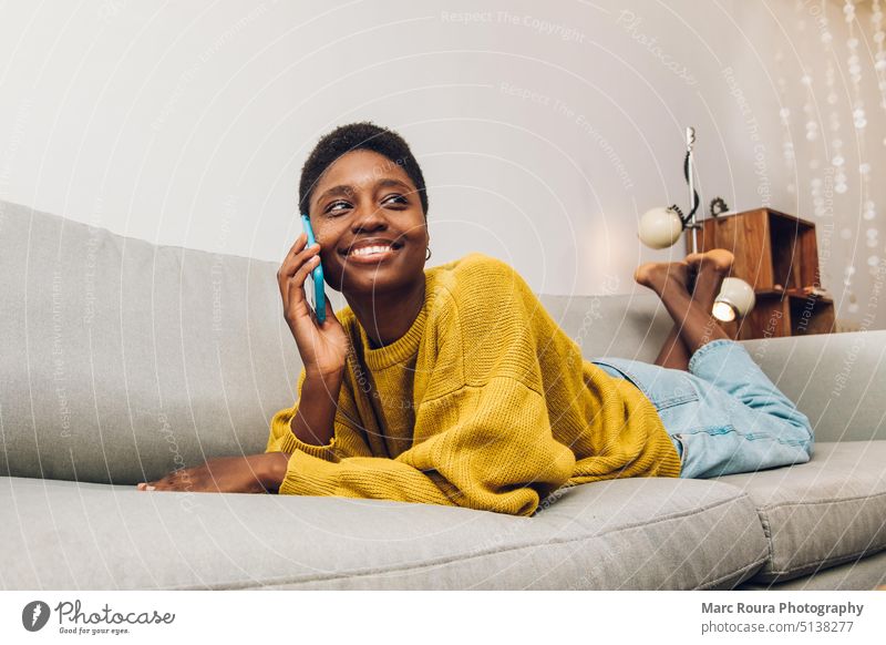Afrikanische Frau, die zu Hause auf dem Sofa sitzend ein Mobiltelefon benutzt Erwachsener Business Anruf lässig Funktelefon Talkrunde heiter Komfort Mitteilung