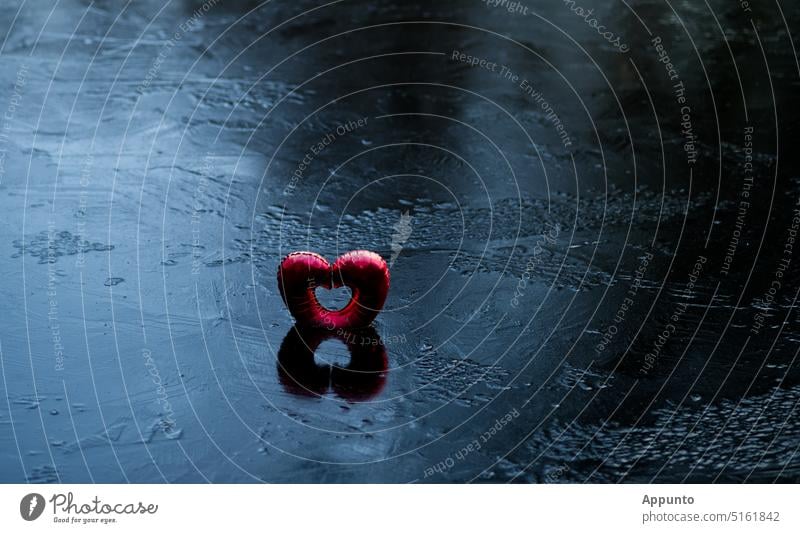 "Liebe gefangen im Eis" (Ein roter herzförmiger Luftballon steht eingefroren in der winterlich vereisten, blauschwarz schimmernden Wasseroberfläche eines Sees)