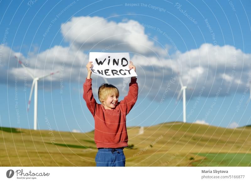 Öko-Aktivist Junge mit Banner "Windenergie" auf dem Hintergrund von Kraftwerken für erneuerbare elektrische Energieerzeugung. Kind und Windmühlen. Windturbinen zur Stromerzeugung. Grüne Energie