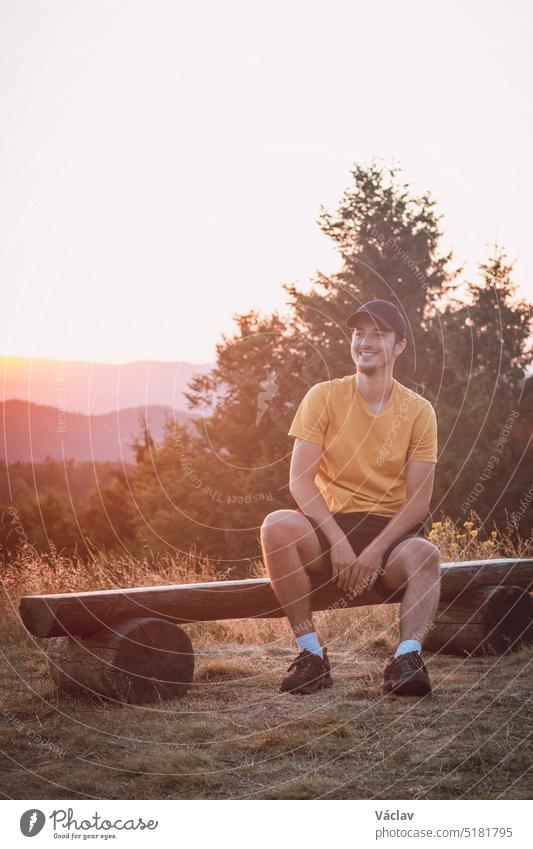 Ein begeisterter Reisender sitzt lächelnd auf einer Holzbank und beobachtet die letzten Reste der Sonne in der rauen Natur. Sonnenuntergang am Horni Becva, Beskiden, Tschechische Republik