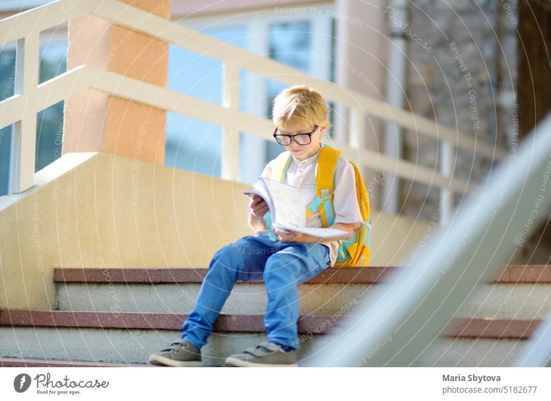 Kluges kleines Kind sitzt und liest auf der Treppe eines Schulgebäudes. Hochwertige Bildung für Kinder. Porträt eines lustigen Streber-Schuljungen mit großer Brille. Vision Probleme. Zurück zu Schule Konzept.