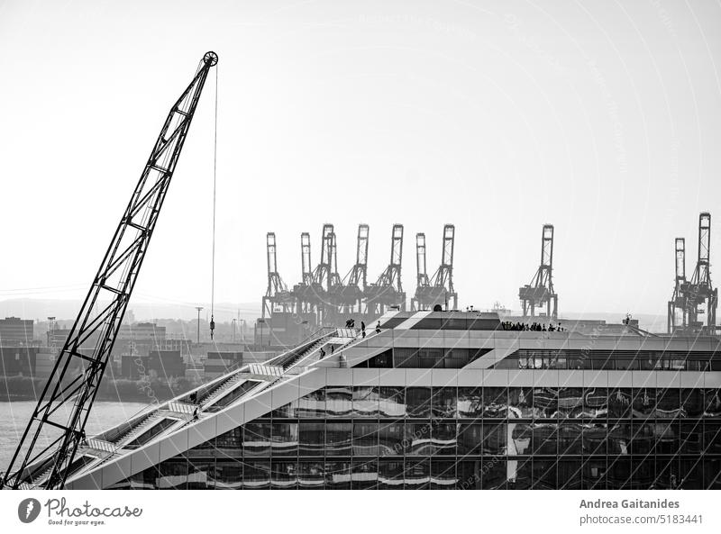 Seitansicht auf einen Teil vom Dockland mit Personen auf der Treppe und der Aussichtsplattform, im Hintergrund Hafenkräne und Container, schwarz-weiß, horizontal