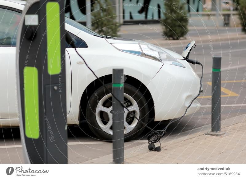 Aufladen moderner Elektroautos, New Energy Vehicles, NEV, auf der Straße weißes Elektroauto mit angeschlossenem Kabel und einer Ladestation. Umweltfreundliches alternatives Energiekonzept.