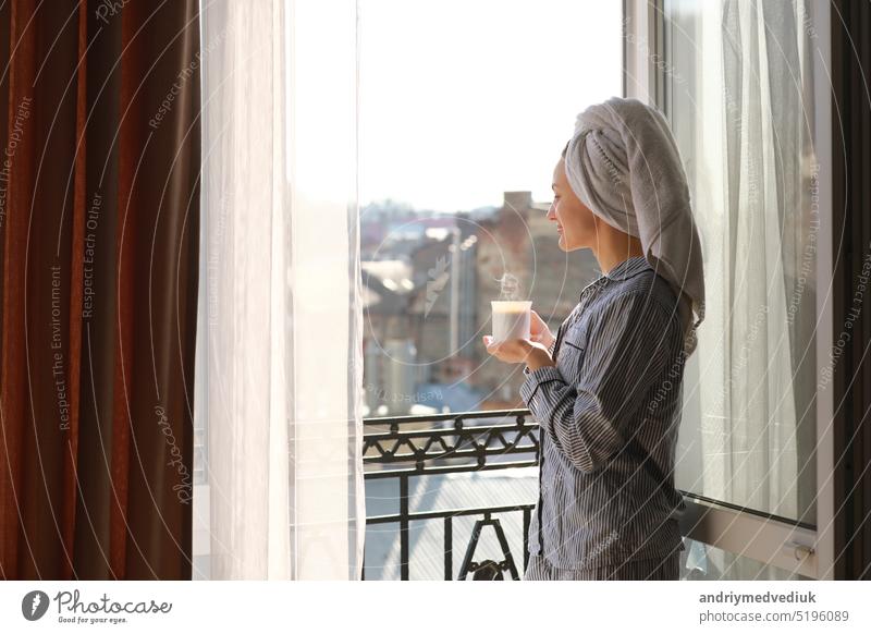 Lächelnde junge Frau in Nachtwäsche und Handtuch auf dem Kopf hält eine Tasse heißen Kaffee oder Tee auf dem Balkon am Fenster am Morgen. Konzept des häuslichen Lebensstils.