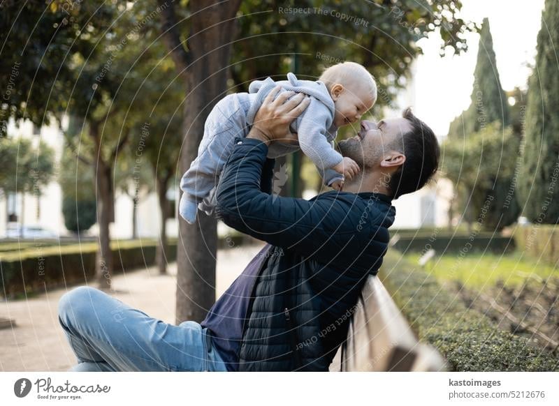 Vater hält und hebt seine niedlichen Säugling Baby Junge Kind sitzt auf Holzbank in städtischen Stadtpark. Vater und Sohn genießen einen unberührten Moment des Glücks, lächelnd und lachend.