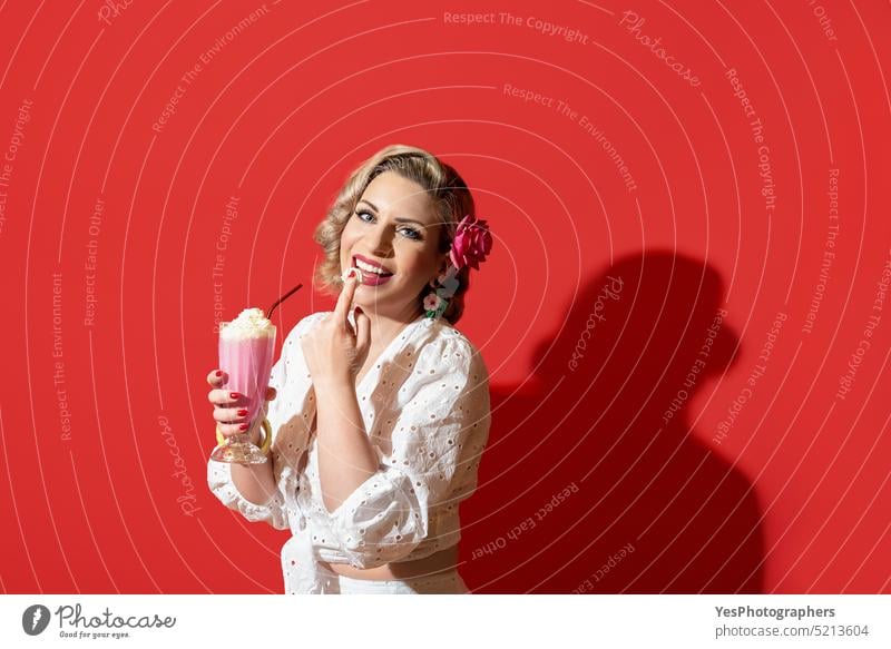 Frau trinkt einen Milchshake vor einem roten Hintergrund. Retro-Stil Frau Porträt 50s Schönheit blond blaue Augen Farbe Konzept Sahne lockig kurvenreich