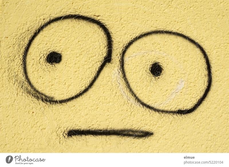 an eine gelbe Wand mit schwarz gezeichnetes Emoji mit großen Augen und neutralem Ausdruck Fratze Grimasse Smiley Smiley-Zeichnung neutraler Blick zeichnen