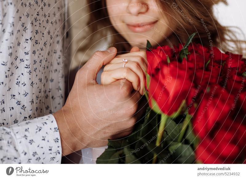 Junger Mann hält die Hand seiner Verlobten mit goldenem Ring und macht ihr einen Heiratsantrag mit einem Strauß roter Rosen. Verlobung eines jungen verliebten Paares. Das Konzept der Liebe und Zweisamkeit.