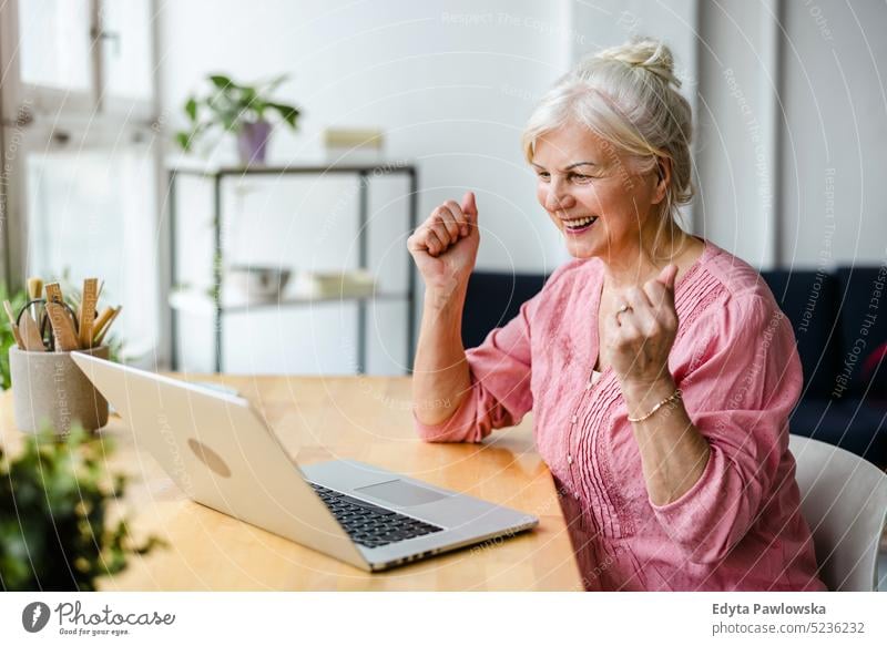 Fröhliche ältere Frau, die im heimischen Wohnzimmer einen Laptop benutzt echte Menschen Senior im Innenbereich Loft Fenster heimwärts reifer Erwachsener