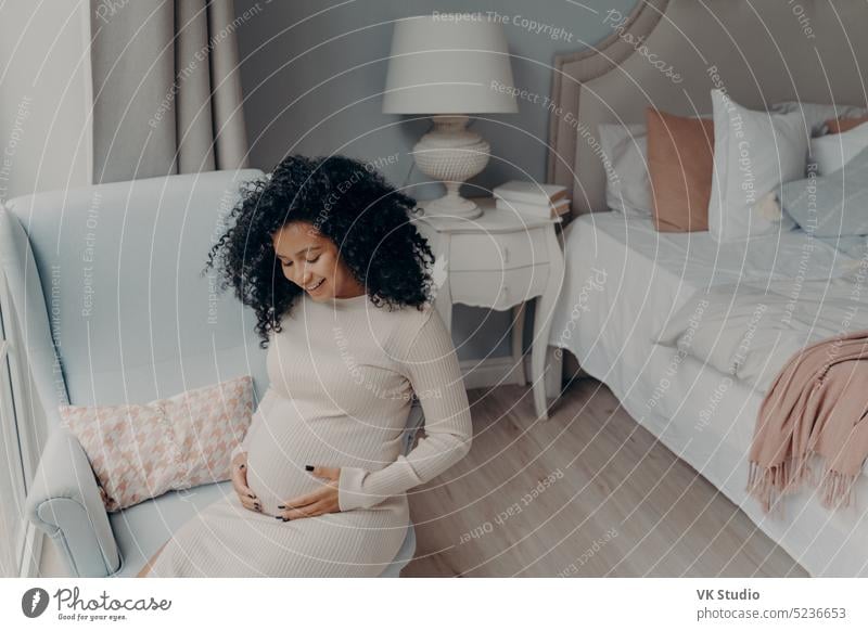 Hübsche Afroamerikanerin genießt die glückliche Schwangerschaft, während sie im Schlafzimmer sitzt schwanger Afrikanisch gemischte Rasse jung Glück Frau Baby