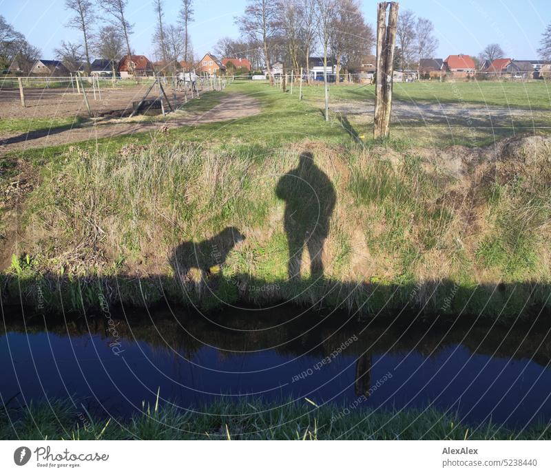 Schattenspiel auf einer Wiese am Wassergraben, welches einen Hund und einen Mann zeigt, im Hintergrund eine Koppel und Häuser eines Dorfes, Bäume, Gras