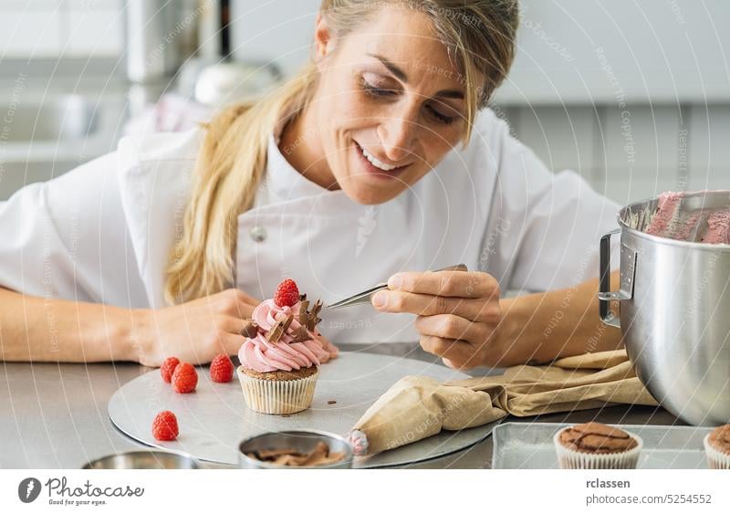 Frauen in einer Konditorei, die mit einer Pinzette Schokoladenstückchen auf einem Muffinbelag drapieren Spritzbeutel rosa Himbeeren Kuchen Cupcake Geburtstag
