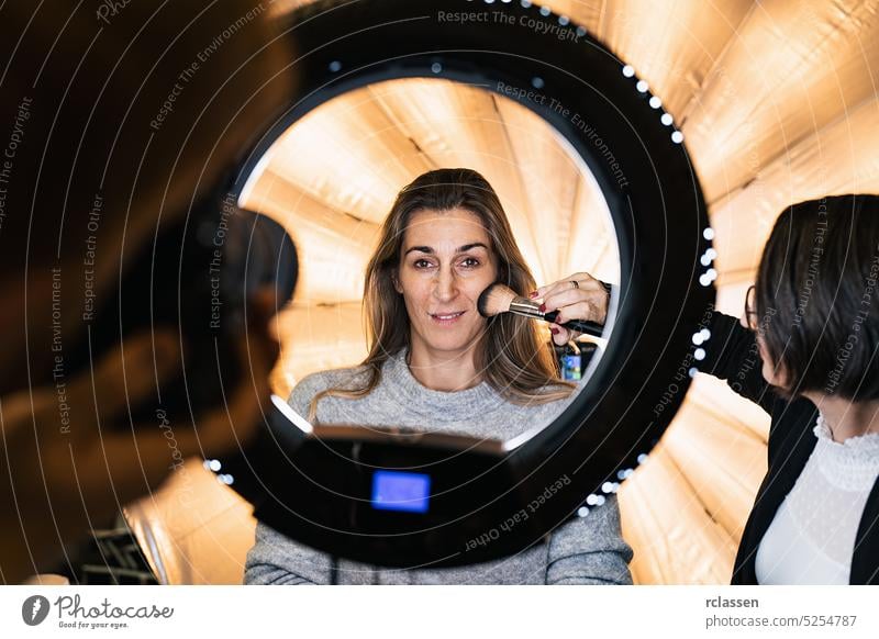 Maskenbildnerin pudert das Gesicht eines Models, das vor der Kamera durch eine Ringlichtlampe posiert, bei einem Fotoshooting mit Ringlicht Puderpinsel