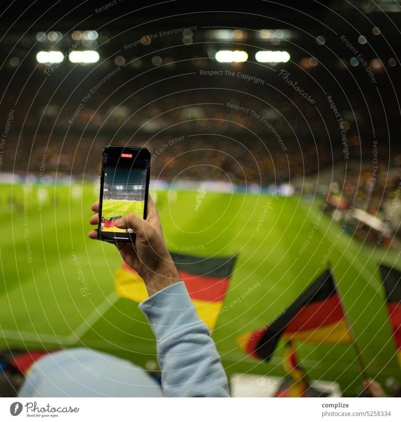 Ein Fussball Fan filmt mit seinem Smartphone ein Fussball-Spiel im Stadion mit wehenden deutschen Flaggen zur EM oder WM Fans filmen Fotografie Fahne