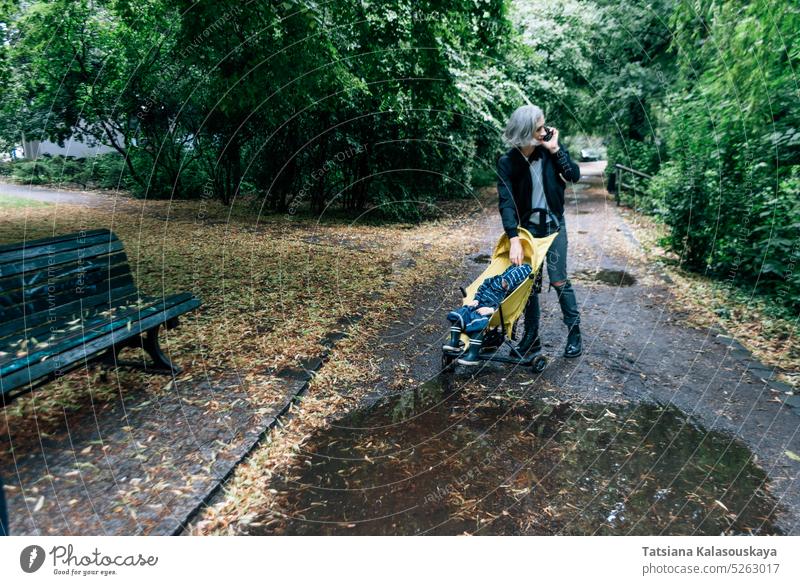 Eine Frau geht mit einem Kinderwagen im Park spazieren und spricht am Telefon Junge Sohn Mutter Mutterschaft Mama Kindererziehung kaukasische Ethnizität