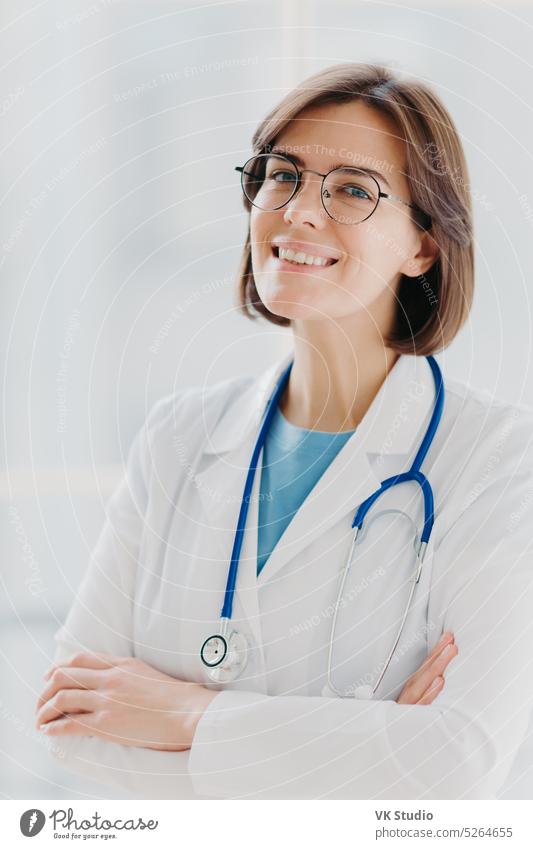 Close up Porträt von kurzhaarigen weiblichen Allgemeinmediziner steht mit Lächeln und Arme verschränkt, verwendet Stethoskop, genießt die Arbeit, posiert gegen weißen Hintergrund. Menschen, medizinische Versorgung und Behandlung Konzept