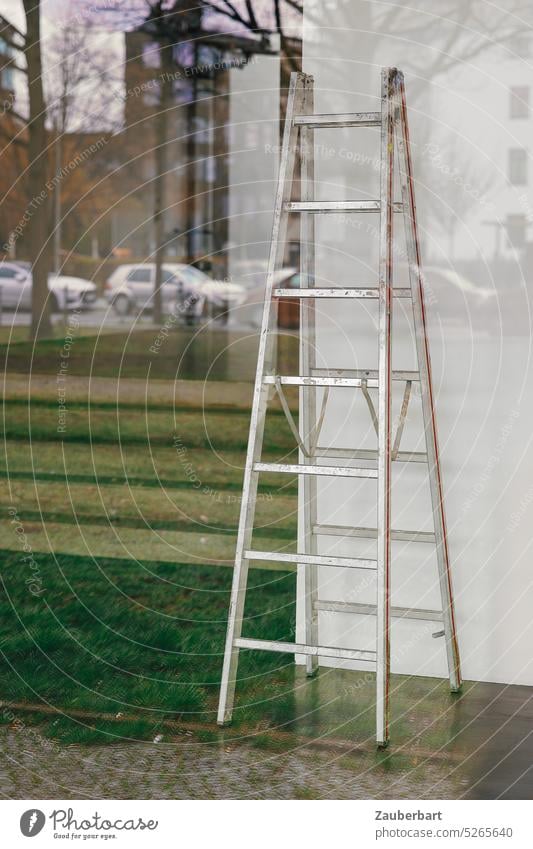 Leiter in Ladengeschäft hinter spiegelnder Scheibe, darauf Wiese und städtische Bebauung Spiegelung Schaufenster Baustelle Aufstieg einrichten Einrichtung