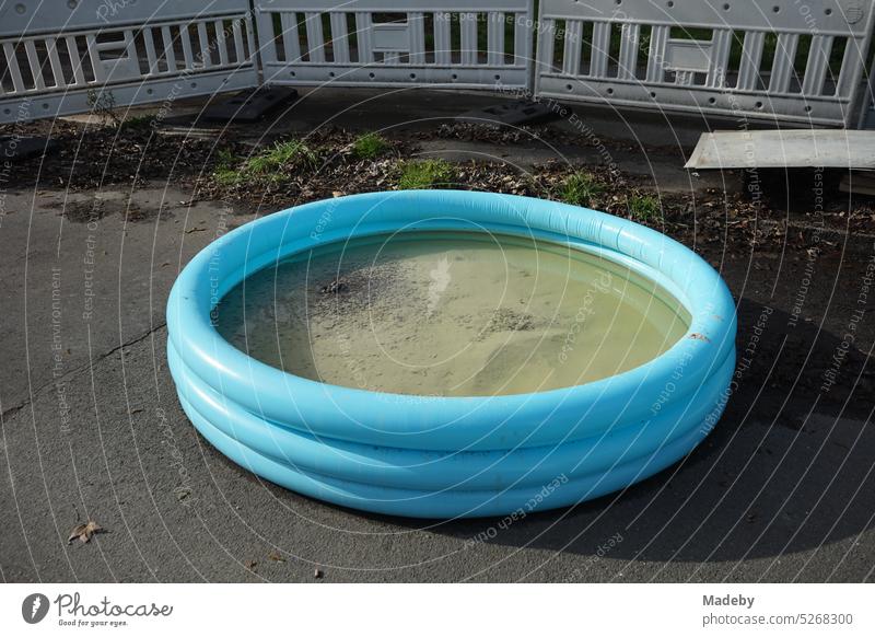 Aufgeblaseneres blaues rundes Planschbecken mit Sand und Wasser auf grauem Asphalt auf einer Baustelle mit Absperrung im Frühling bei Sonnenschein in der Innenstadt von Wuppertal in Nordrhein-Westfalen
