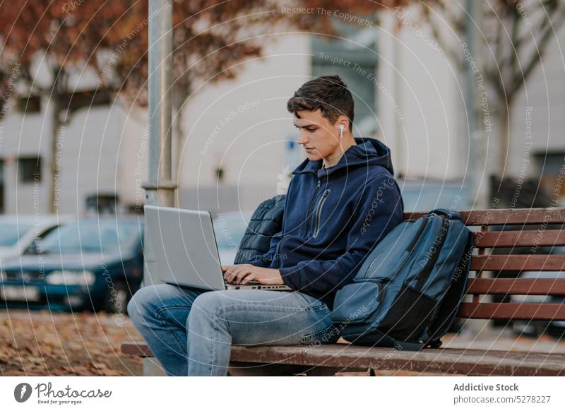 Konzentrierter Mann, der auf einer Bank am Laptop sitzt Kopfhörer Park Schüler Herbst Fokus Konzentration fallen jung männlich warme Kleidung Apparatur Browsen