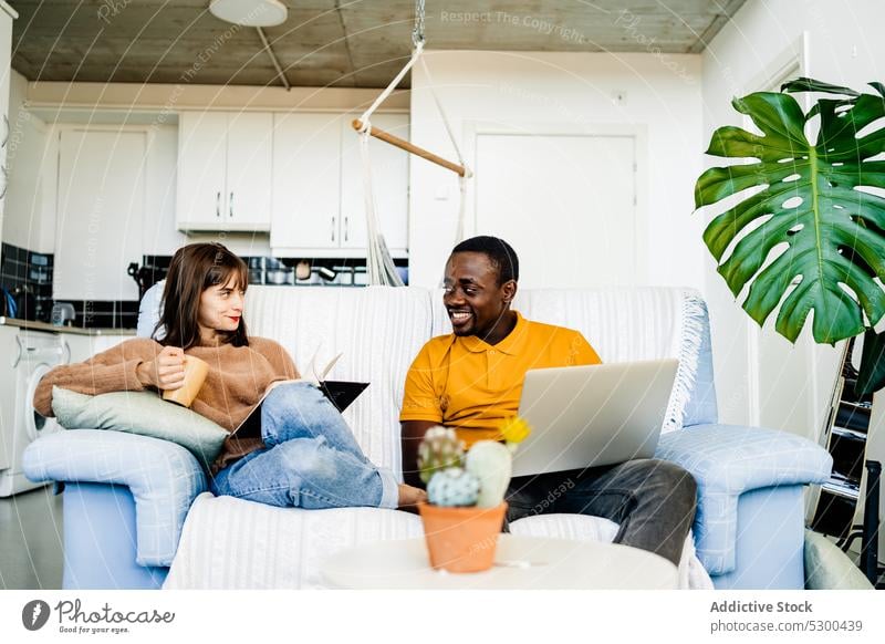 Fröhliches Paar mit Buch und Laptop zu Hause lesen benutzend Sofa Glück Lächeln heiter Liebe Zusammensein Partnerschaft Wohnzimmer multiethnisch heimwärts
