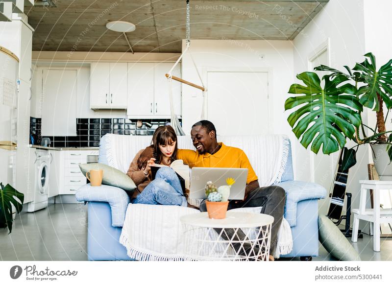 Fröhliches Paar mit Buch und Laptop zu Hause lesen benutzend Sofa Glück Lächeln heiter Liebe Zusammensein Partnerschaft Wohnzimmer multiethnisch heimwärts