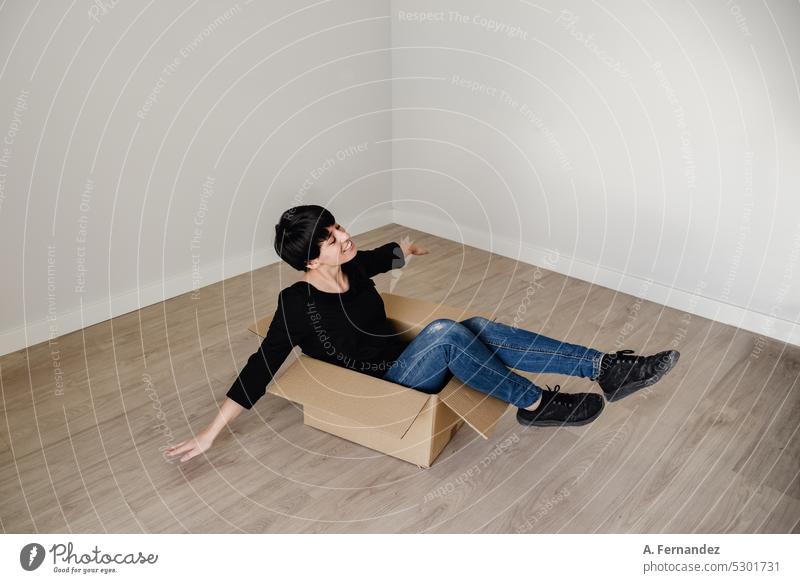 Eine junge Frau mit kurzen Haaren in einem Karton in einem leeren Raum. Konzept des Auszugs und des Beginns eines neuen Lebens. Spielen mit Kartons, kreative Phantasie