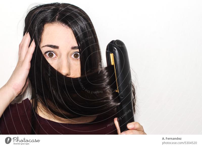 Detail einer Frau mit langen Haaren und einer Geste der Überraschung bei der Verwendung eines Haarglätters auf weißem Hintergrund. Konzept der Verwendung von Wärme zum Glätten der Haare.