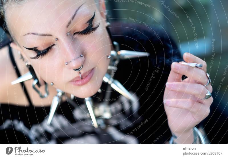 Porträt eines jungen sexy, verführerischen Gothic-Mädchens mit Piercings, das ein großes Stachelhalsband trägt, mit geschlossenen Augen und aufgeklebten künstlichen Wimpern, das Zigarette raucht