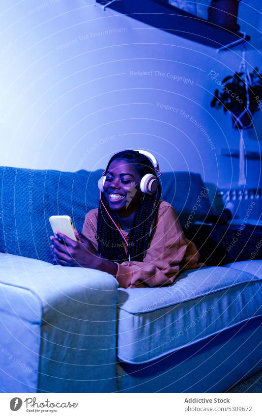 Glückliche schwarze Frau, die mit ihrem Smartphone Nachrichten schreibt und auf dem Sofa Musik hört Kopfhörer benutzend Lügen Gesang Browsen neonfarbig heiter