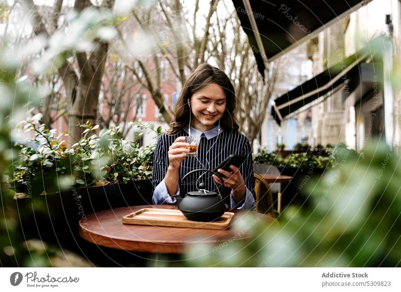 Frau benutzt Smarthone auf der Terrasse eines Cafés Smartphone positiv Kopfhörer zuschauen benutzend Tee Teekanne Handy online Internet Gerät Apparatur