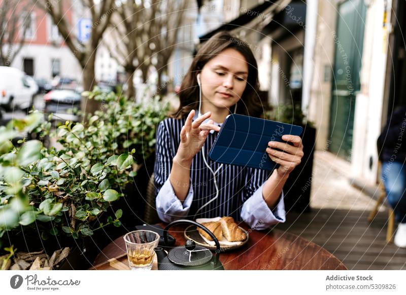 Frau mit Tablet auf der Terrasse eines Cafés positiv Tablette Kopfhörer zuschauen benutzend Lebensmittel Tee Teekanne Croissant online Internet Gerät Apparatur