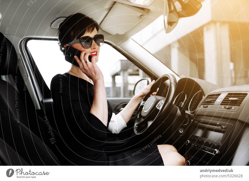 Attraktive erfolgreiche Geschäftsfrau sitzt im Inneren des Luxus-Auto und sprechen auf ihrem Smartphone. Geschäftsinhaberin Frau mit Sonnenbrille fahren ein Auto löst Arbeit Fragen auf dem Weg ins Büro. Lebensstil
