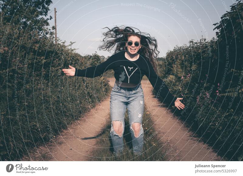 Glückliches Mädchen mit ausgestreckten Armen und Sonnenbrille auf einem Feldweg Schirme gedämpfter Ton Schwarzer Pullover Arme ausgestreckt Löcher in Jeans