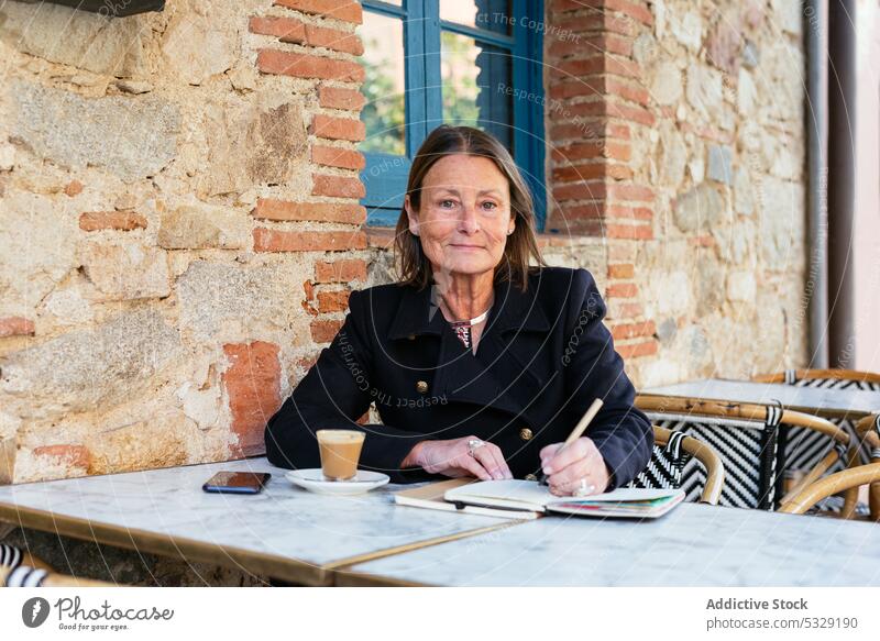 Fröhliche Frau mit Notizbuch am Tisch sitzend schreiben zur Kenntnis nehmen Café positiv Lächeln Planer Glück heiter Notebook Hinweis Kaffee Tasse Arbeit Straße