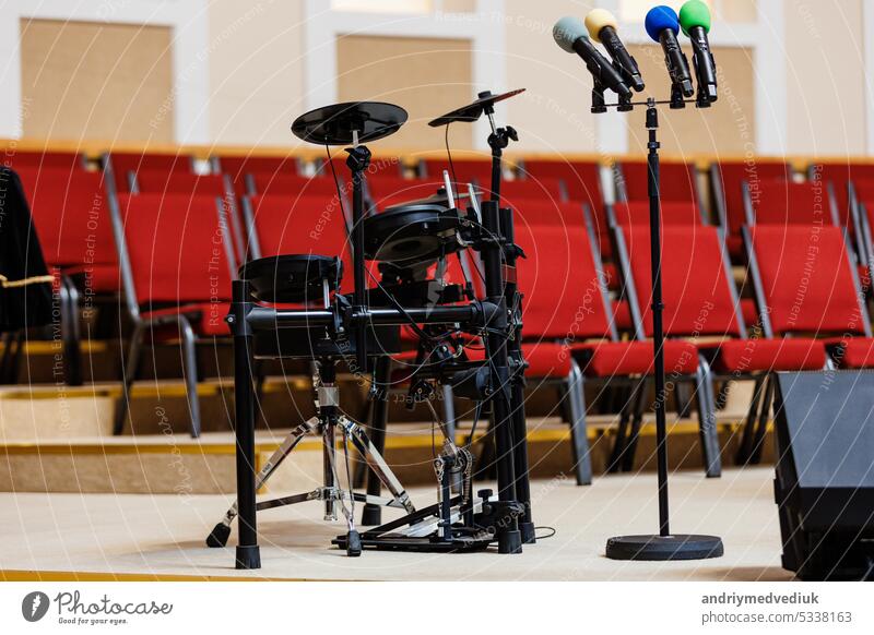 Bunte Mikrofone auf einem Ständer und ein elektronisches Schlagzeug auf einem Podium, viele rote Stühle verschwommen im Hintergrund. Konzept für Konzert, Veranstaltung, Kommunikation, Seminar, Medien, Abschlussfeier, Performance
