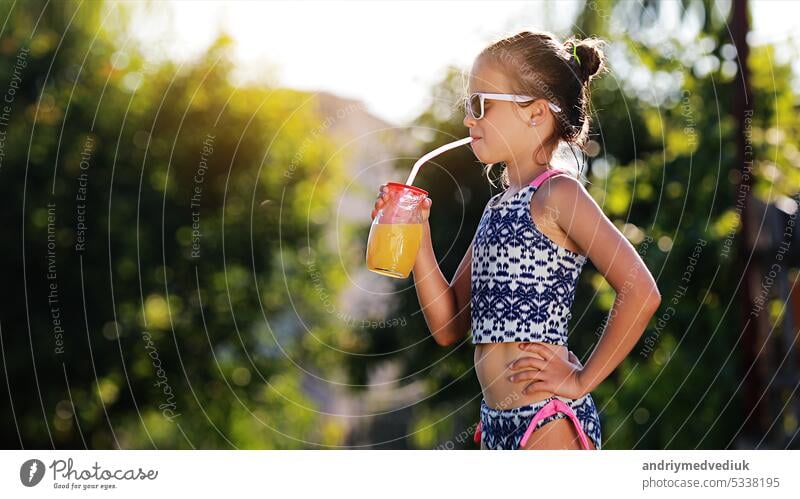 Schönes kleines Mädchen, süßes Kleinkind mit Sonnenbrille trinkt Orangensaft und zeigt Daumen nach oben. Kid in in Badebekleidung Spaß während Familienurlaub in einem Resort in der Nähe von Pool. Sommerferien