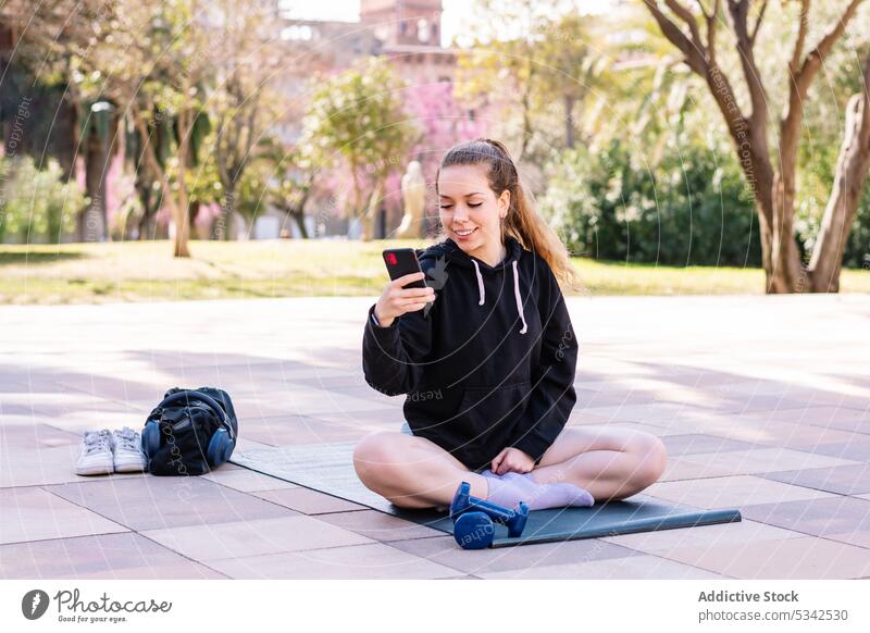 Fröhliche junge Frau benutzt ihr Smartphone beim Yoga benutzend online meditieren sukhasana üben Wellness Browsen Internet Straße sich[Akk] entspannen Asana