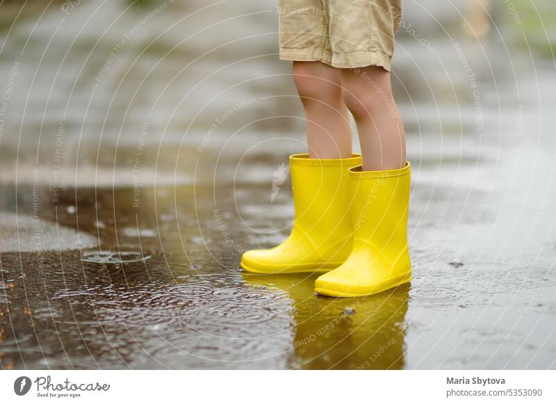 Kleiner Junge in gelben Gummistiefeln, der an einem regnerischen Sommertag in einer kleinen Stadt spazieren geht. Kind hat Spaß. Draußen Spiele für Kinder in regen.
