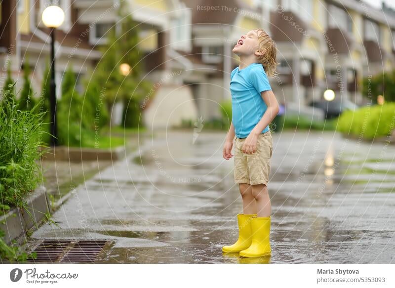 Kleiner Junge in gelben Gummistiefeln, der an einem regnerischen Sommertag in einer kleinen Stadt spazieren geht. Kind hat Spaß. Kind fängt Regentropfen mit Zunge. Draußen Spiele für Kinder in regen.
