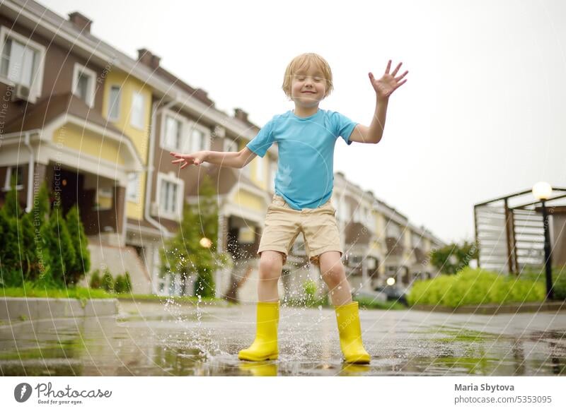 Kleiner Junge in gelben Gummistiefeln, der an einem regnerischen Sommertag in einer Kleinstadt in eine Wasserpfütze springt. Kind hat Spaß. Draußen Spiele für Kinder in regen.