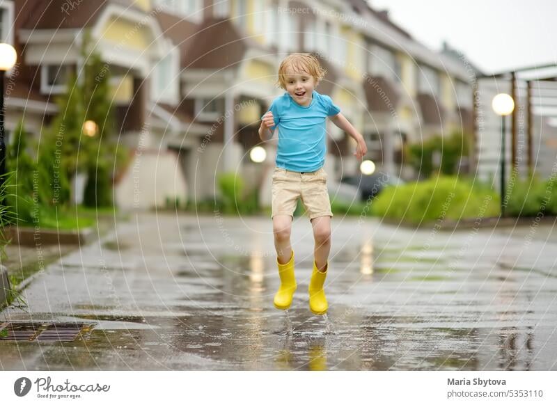 Kleiner Junge in gelben Gummistiefeln, der an einem regnerischen Sommertag in einer Kleinstadt in eine Wasserpfütze springt. Kind hat Spaß. Draußen Spiele für Kinder in regen.