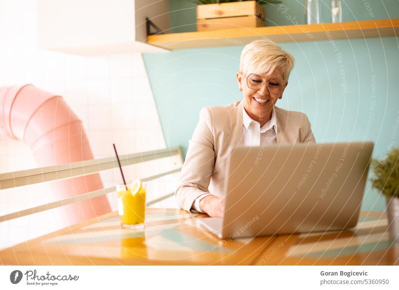 Ältere Frau, die ein Mobiltelefon benutzt, während sie am Laptop arbeitet und frischen Orangensaft im Café trinkt gealtert Anruf Kaffee Kaffeehaus Mitteilung