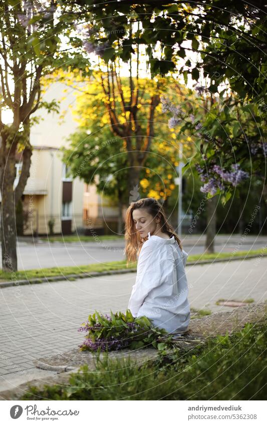 junges schönes Mädchen in einem weißen Hemd und schwarzer Hose sitzt in der Nähe von blühenden Fliederbäumen mit lila Blumen im Freien, blühenden Baum, Frühling, Sonne, Glück, Zärtlichkeit.