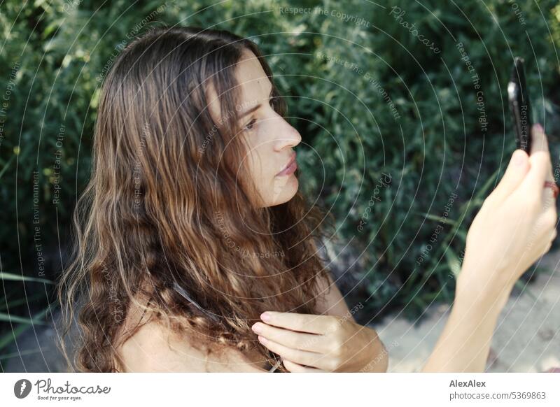 Portrait einer jungen, brünetten, langhaarigen Frau, ihr Smartphone als Spiegel benutzt   anschaut am Strand vor buschigen Pflanzen junge Frau Locken schön fit