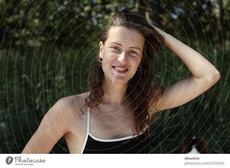 Portrait einer jungen, schönen, brünetten, lächelnden Frau, die im Sand vor einem Wald sitzt warm sommerlich schönes wetter Sommer Pflanzen Nähe Tag Ruhe