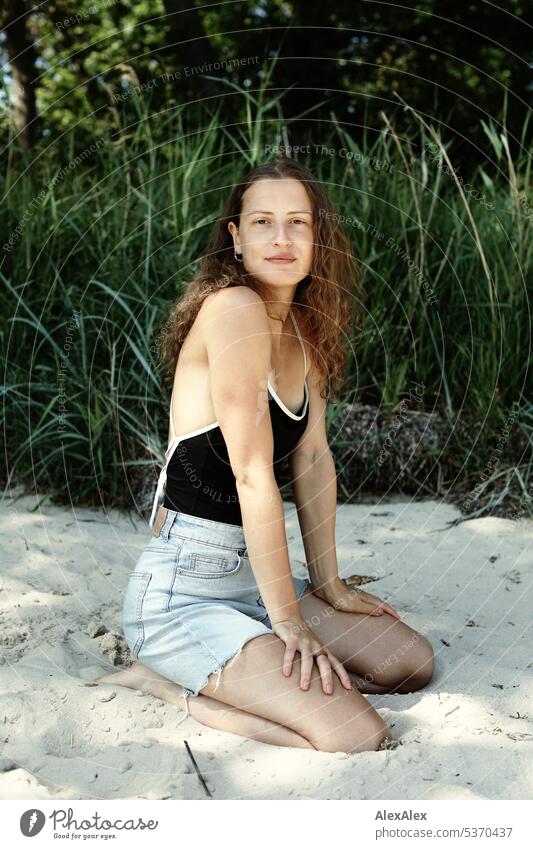 seitliches Portrait einer jungen, schönen, brünetten Frau, die im Sand vor einem Wald kniet warm sommerlich schönes wetter Sommer Pflanzen Nähe Tag Ruhe