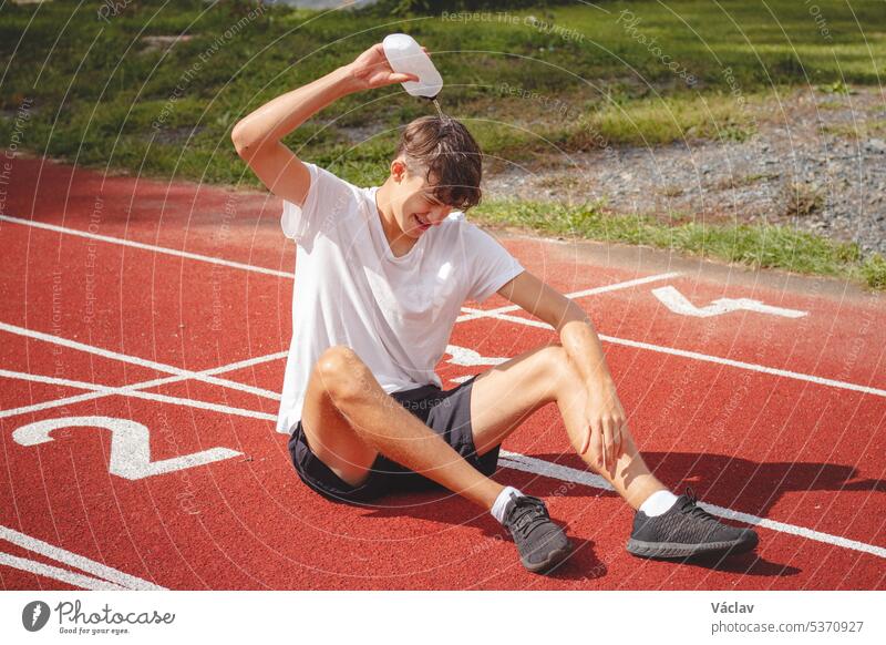 Ein junger Sportler erfrischt sich nach einem harten Training auf dem Leichtathletik-Oval in der großen Hitze mit Wasser. Ausdauertraining. Braunhaariger Jugendlicher