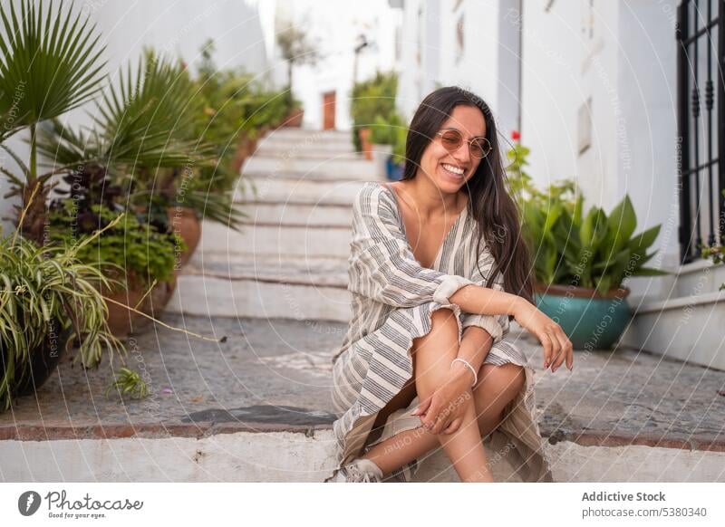 Lächelnde Frau sitzt auf den Stufen einer Straße Treppe Gebäude Stadt Pflanze Hand am Kinn froh positiv jung Glück heiter Sonnenbrille eingetopft ruhen Stil
