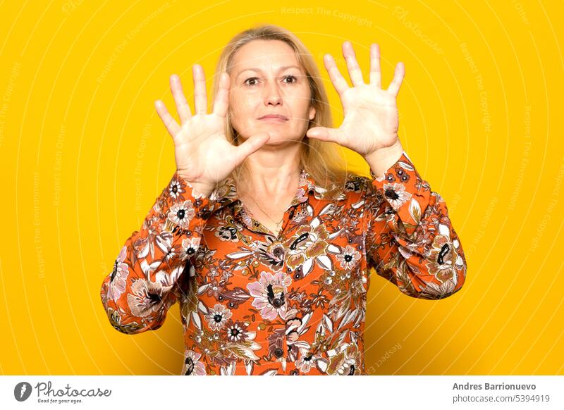 Kaukasische blonde Frau in den 40ern, die einen gemusterten Pullover trägt und besorgt mit ihren Handflächen vor ihrem Gesicht posiert, isoliert auf gelbem Studiohintergrund.