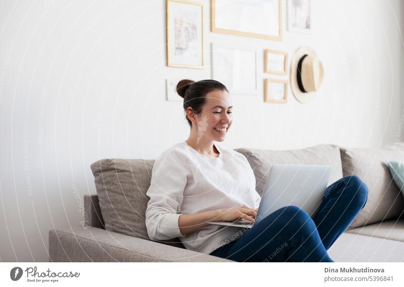Junge Frau arbeitet am Laptop. Millennial Mädchen arbeiten Remote-Job von zu Hause, mit Handy, sitzen auf dem gemütlichen Sofa, lächelnd. Hintergrund schön